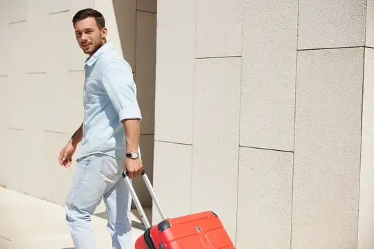 Молодой человек в повседневной одежде идет, держа в руках красный чемодан, и оглядывается через плечо на фоне современного городского пейзажа