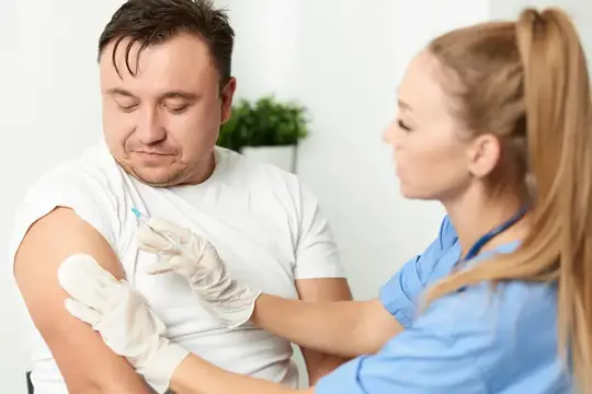 Медсестра делает укол в плечо мужчине в белой футболке