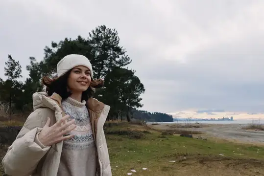 Улыбающаяся девушка в белой шапке и пуховике на фоне залива