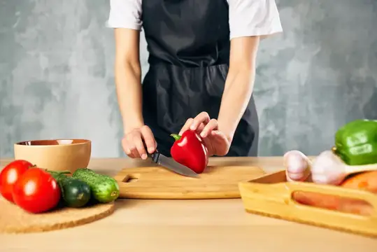 Повар в чёрном фартуке режет красный перец на доске