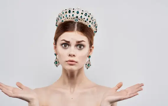 Удивлённая женщина в короне с изумрудами разводит руками