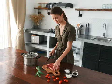 Женщина на кухне улыбается, готовит ужин из мяса и овощей на деревянном столе на фоне стильной кухни, образ жизни