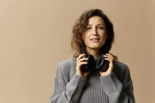 Веселая, радостная, симпатичная кудрявая красавица в сером повседневном свитере в наушниках смотрит в камеру, позируя изолированно на бежевом пастельном фоне