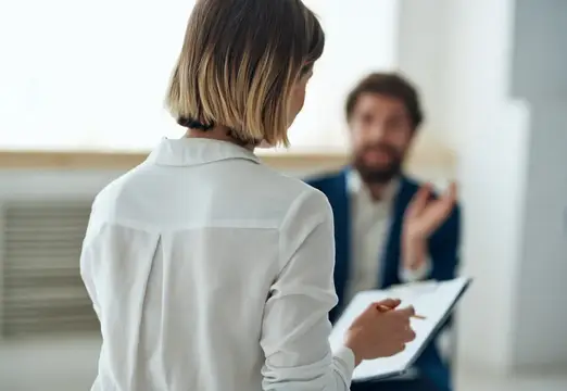 Женщина с блокнотом слушает бородатого мужчину в офисе