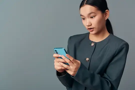 Удивлённая азиатская девушка смотрит в смартфон на сером фоне