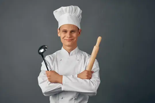 Улыбающийся молодой повар с половником и скалкой на сером фоне