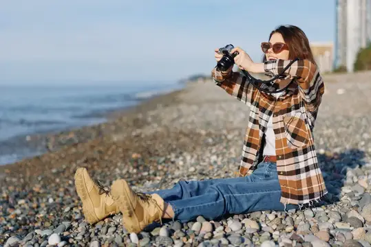 Девушка в клетчатой рубашке фотографирует на каменистом берегу моря