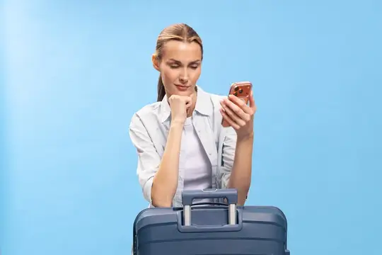 Задумчивая светловолосая женщина с телефоном у чемодана на голубом фоне