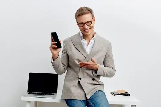 Улыбающийся мужчина в очках смотрит на смартфон, сидя на столе с ноутбуком