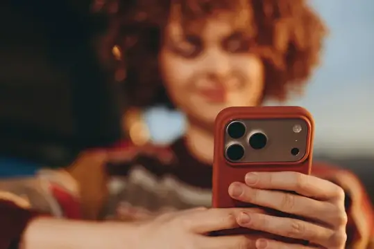 Рыжеволосая девушка держит смартфон в красном чехле на фоне заката