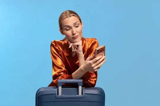 Женщина со светлыми волосами, одетая в оранжевую рубашку, отдыхает, опираясь на чемодан, держит телефон и смотрит на экран