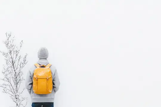 Одинокая фигура в серой толстовке и вязаной шапке стоит на фоне белой стены, демонстрируя зимнее одиночество и спокойствие с ярким желтым рюкзаком, передавая чувство приключения