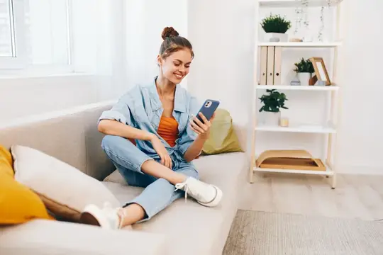 Улыбающаяся девушка сидит на диване со смартфоном в руках