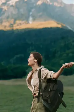 Энергичная женщина в походе, смеется, запрокинув голову, на фоне живописных гор, свежий воздух, яркая зелень, непринужденная атмосфера путешествия, залитый солнцем момент, современный гардероб