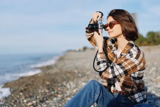 Девушка в клетчатой рубашке фотографирует на плёночный фотоаппарат у моря