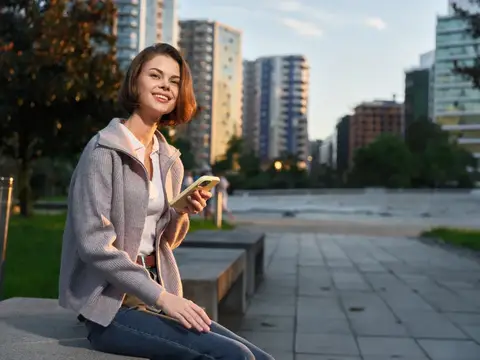 Портрет стильного, расслабленного человека на скамейке возле современных зданий, использующего смартфон, отражающий баланс между природой и городской жизнью в мягком дневном свете