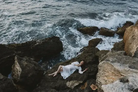 Девушка в белом платье лежит на скалах у бурного моря