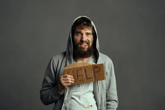 Бородатый мужчина в капюшоне держит картонку с надписью HELP