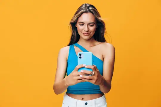 Улыбающаяся девушка в синем топе смотрит в смартфон на жёлтом фоне