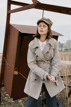 Модная молодая женщина в бежевом тренчкоте и кепке позирует с закрытыми глазами рядом с ржавым металлическим предметом на улице