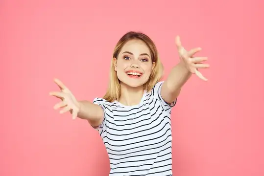 Улыбающаяся блондинка в полосатой футболке тянет руки к камере на розовом фоне