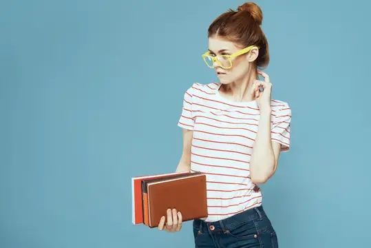 Студентка с книгами в руках на голубом фоне, в жёлтых очках, с модельной причёской, кадрированный вид