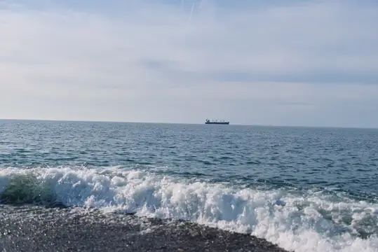 Волна разбивается о гальку на фоне моря и сухогруза