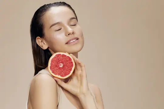 Красивая женщина с грейпфрутом рядом с лицом, уход за чистой кожей и здоровье на бежевом фоне