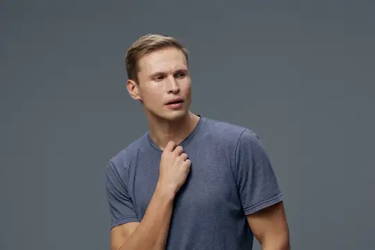 Задумчивый светлокожий мужчина с короткими волосами в серой футболке смотрит в сторону и касается шеи, изолирован на простом сером фоне в студийном портрете