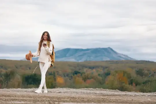Улыбающаяся девушка в белом свитере стоит на фоне осенних гор