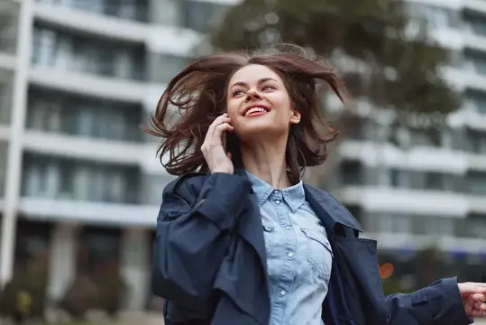 Улыбающаяся женщина в синей куртке пользуется смартфоном на улице, ее волосы развеваются на ветру, она стоит перед современным зданием