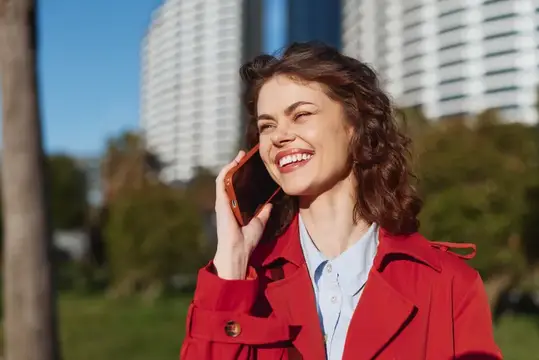 Улыбающаяся темноволосая женщина в красном пальто говорит по телефону на улице