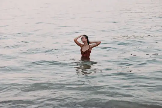 Молодая женщина в бордовом купальнике стоит по грудь в воде и поправляет волосы