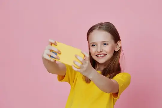 Улыбающаяся девочка снимает на жёлтый смартфон на розовом фоне