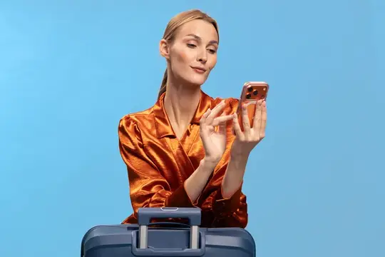Женщина в блестящей оранжевой блузке пользуется смартфоном, рядом с ней стоит синий чемодан