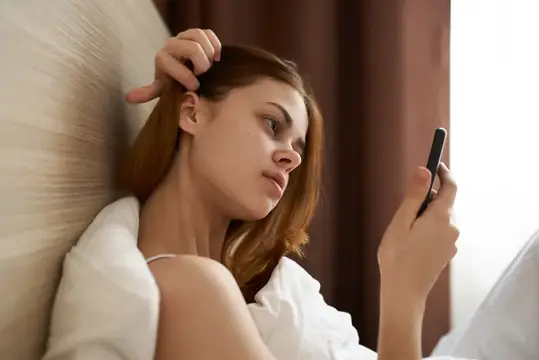 Молодая рыжеволосая женщина лежит в постели и смотрит в смартфон