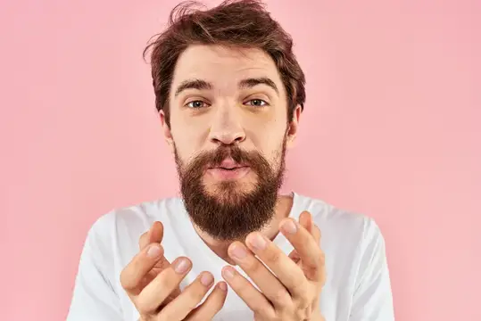 Бородатый мужчина в белой футболке жестикулирует на розовом фоне