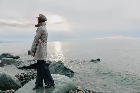 Женщина в плаще и кепке стоит на камнях у моря