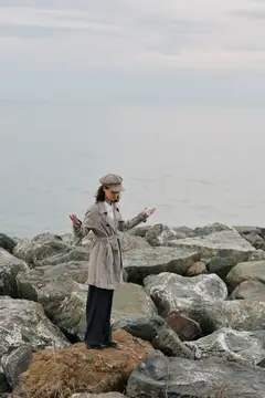 Девушка в плаще и кепке стоит на камнях у моря