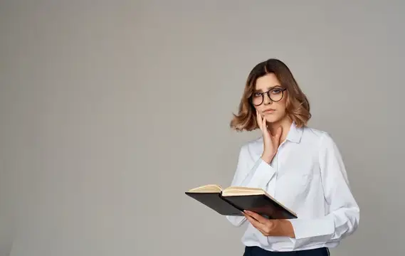 Задумчивая женщина в очках с книгой на сером фоне