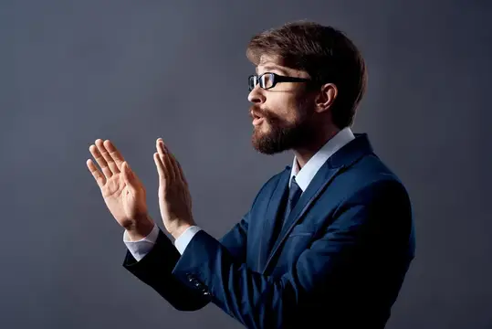Бородатый мужчина в очках и синем костюме жестикулирует руками