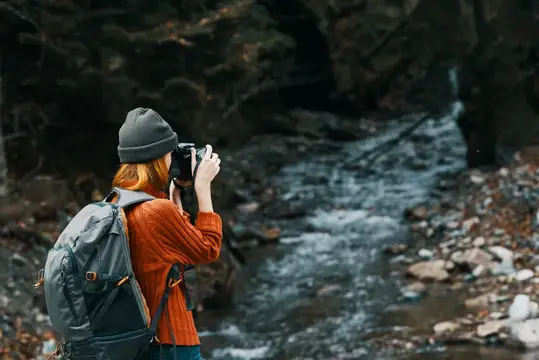 Рыжеволосая девушка с рюкзаком фотографирует горную реку