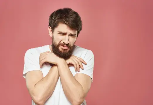 Бородатый мужчина в белой футболке зябко обнимает себя на розовом фоне