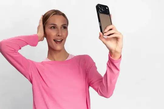 Улыбающаяся женщина в повседневном розовом топе держит смартфон для селфи на однотонном белом фоне