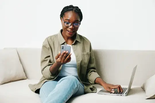 Темнокожая девушка в очках смотрит в смартфон, сидя на диване с ноутбуком