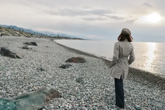 Женщина в плаще стоит на галечном берегу у моря