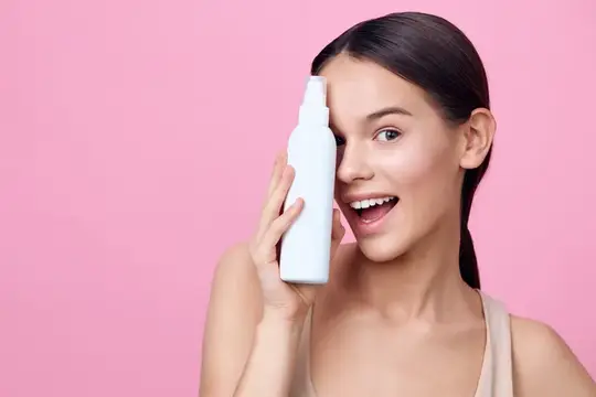 Улыбающаяся темноволосая девушка держит белый флакон на розовом фоне