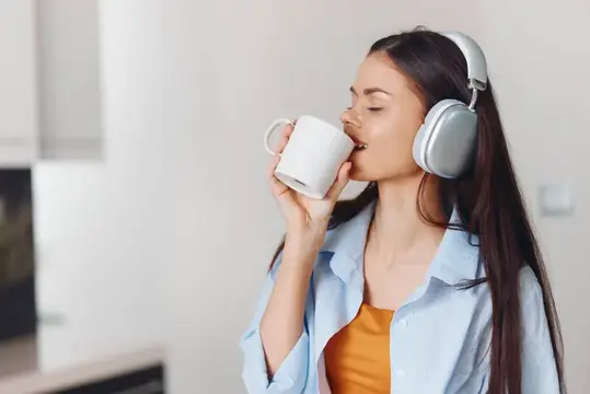 Женщина на кухне в наушниках пьет кофе и слушает музыку на телефоне