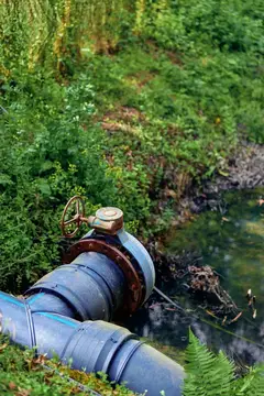 Ржавая труба и вентиль на берегу реки с потоком воды и дренажом, трава и водно-болотная растительность, коррозия металла и инфраструктура на открытом воздухе, требующая обслуживания или ремонта в…