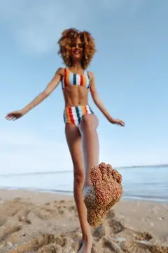 Рыжеволосая девушка в полосатом купальнике весело прыгает на пляже